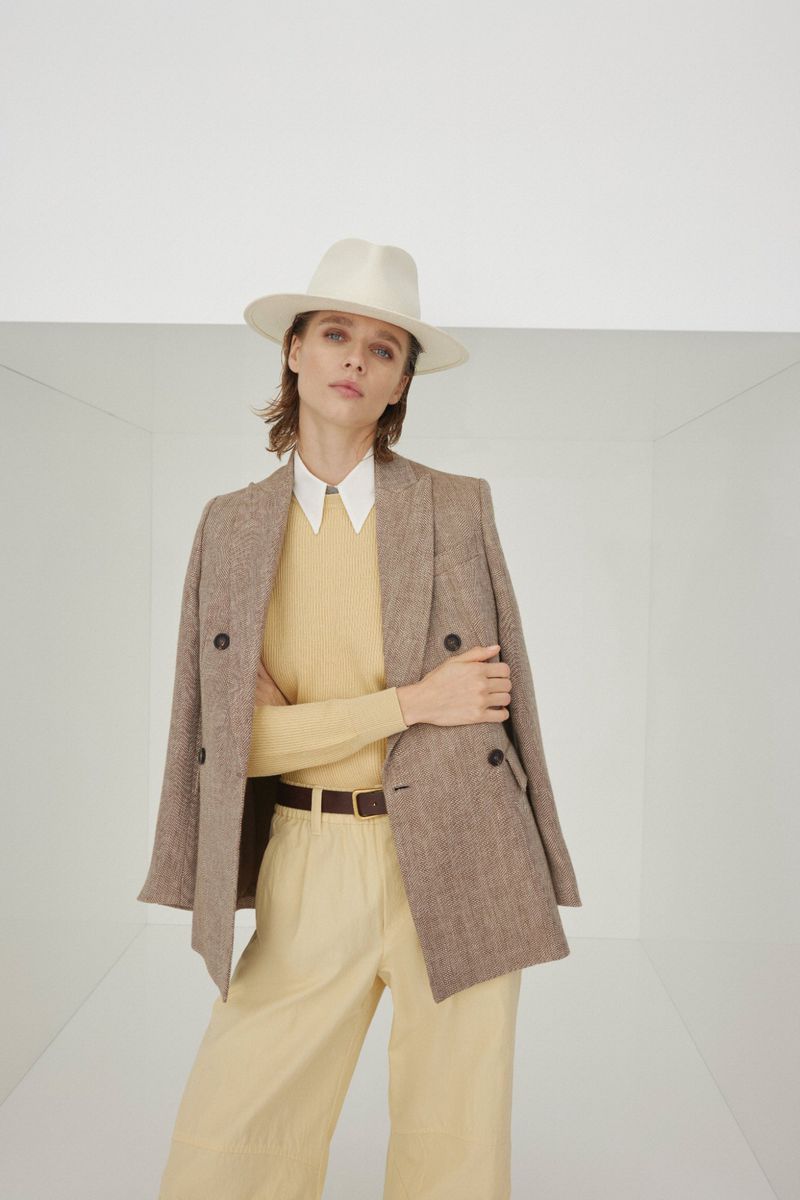 Model wearing a white fedora hat, brown tweed blazer, and butter yellow trousers in a minimalist white studio setting.