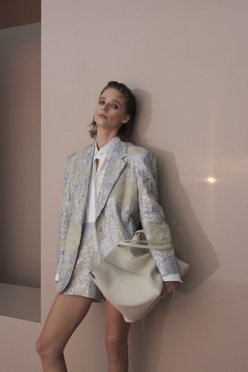 A fashionable woman poses in a patterned blazer and shorts, holding a handbag against a muted studio backdrop with soft, diffused lighting.