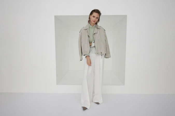 Model wearing a light grey suede jacket over white trousers standing in a minimalist studio setting.