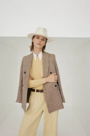 Model wearing a white fedora hat, brown tweed blazer, and butter yellow trousers in a minimalist white studio setting.