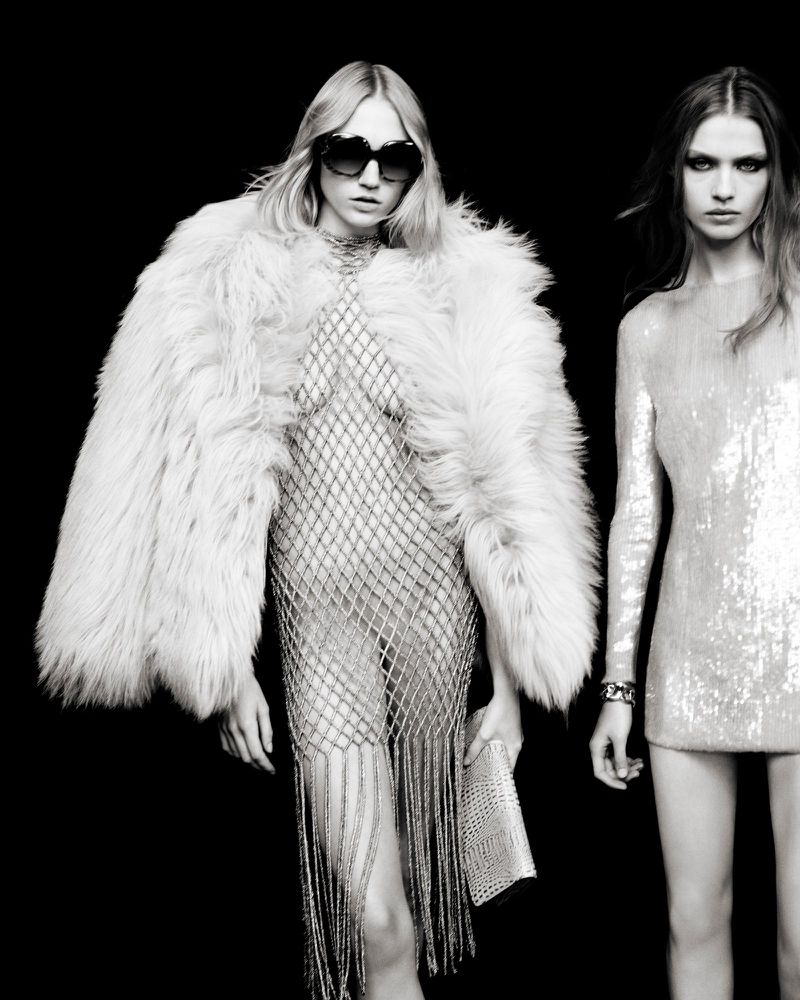 Two fashion models posing in a studio against a dark backdrop, wearing dresses and a fur coat, holding a clutch and wearing sunglasses.
