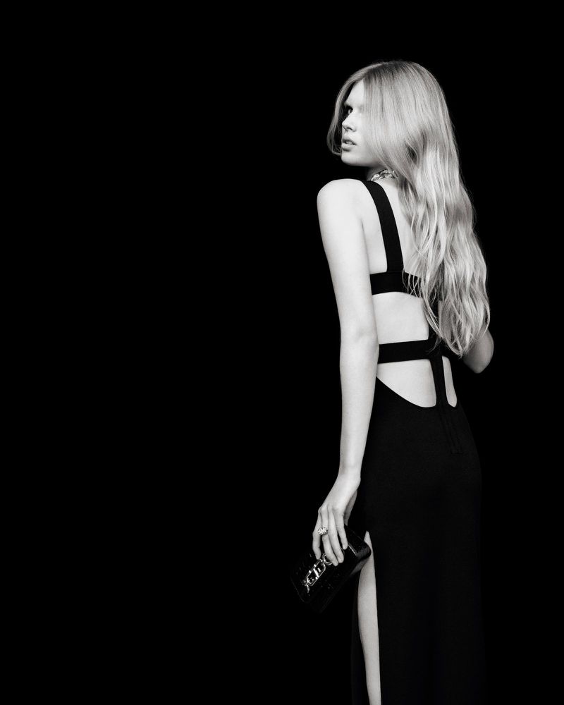Blonde woman in black gown with open back and thigh slit looks over her shoulder while holding a clutch against black background.