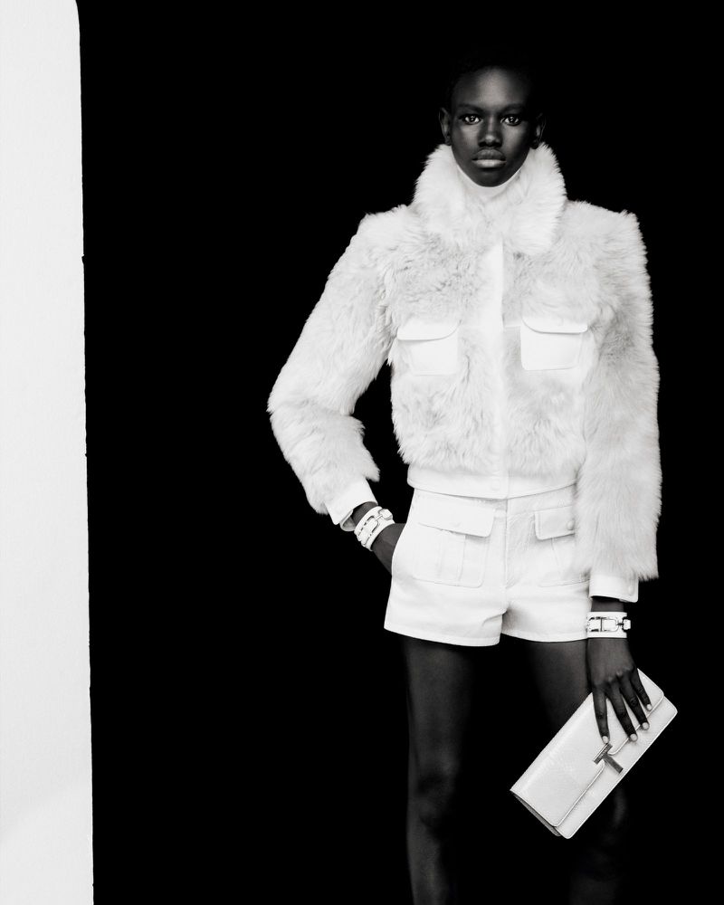 A Black female model in a white faux fur jacket, white shorts, and a clutch poses in front of a solid black backdrop in a studio shot.
