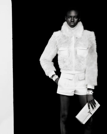 A Black female model in a white faux fur jacket, white shorts, and a clutch poses in front of a solid black backdrop in a studio shot.