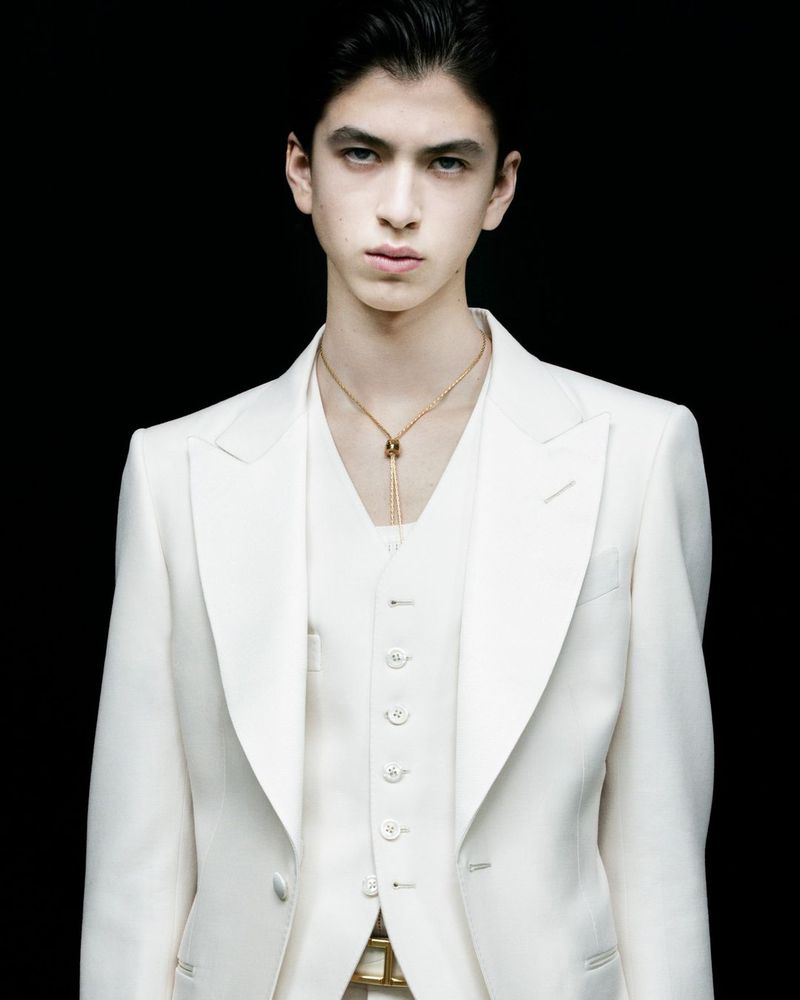 A young man in a light-colored suit stands against a dark background, showcasing a formal, elegant attire with a gold necklace.