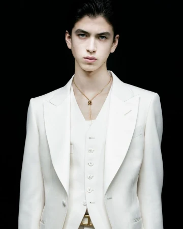 A young man in a light-colored suit stands against a dark background, showcasing a formal, elegant attire with a gold necklace.