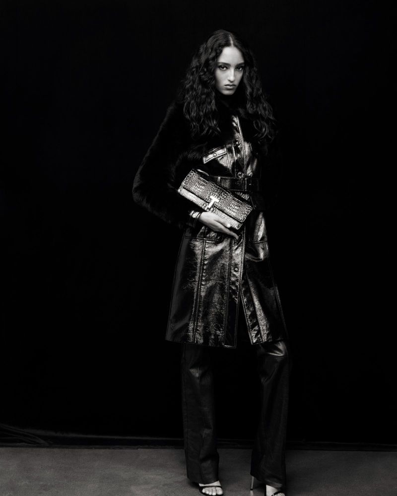 A fashion shot in black and white featuring a female model wearing a shiny black coat, pants, and heels, while holding a handbag.