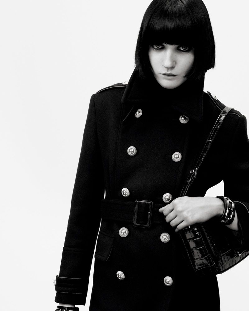 Fashion shot of a woman with dark hair wearing a black belted coat and carrying a black handbag, against a white background.