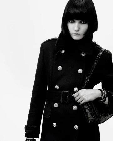 Fashion shot of a woman with dark hair wearing a black belted coat and carrying a black handbag, against a white background.