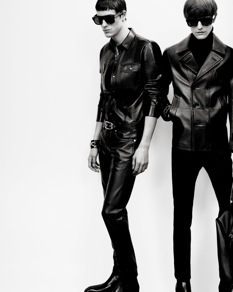 Two men model black leather outfits in a stark, high-contrast fashion photograph with dramatic lighting against a white background.