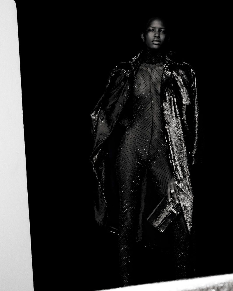 A Black woman stands in a dark studio wearing a shimmery catsuit and coat, holding a transparent rectangular clutch, with a neutral expression.