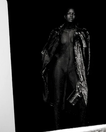 A Black woman stands in a dark studio wearing a shimmery catsuit and coat, holding a transparent rectangular clutch, with a neutral expression.