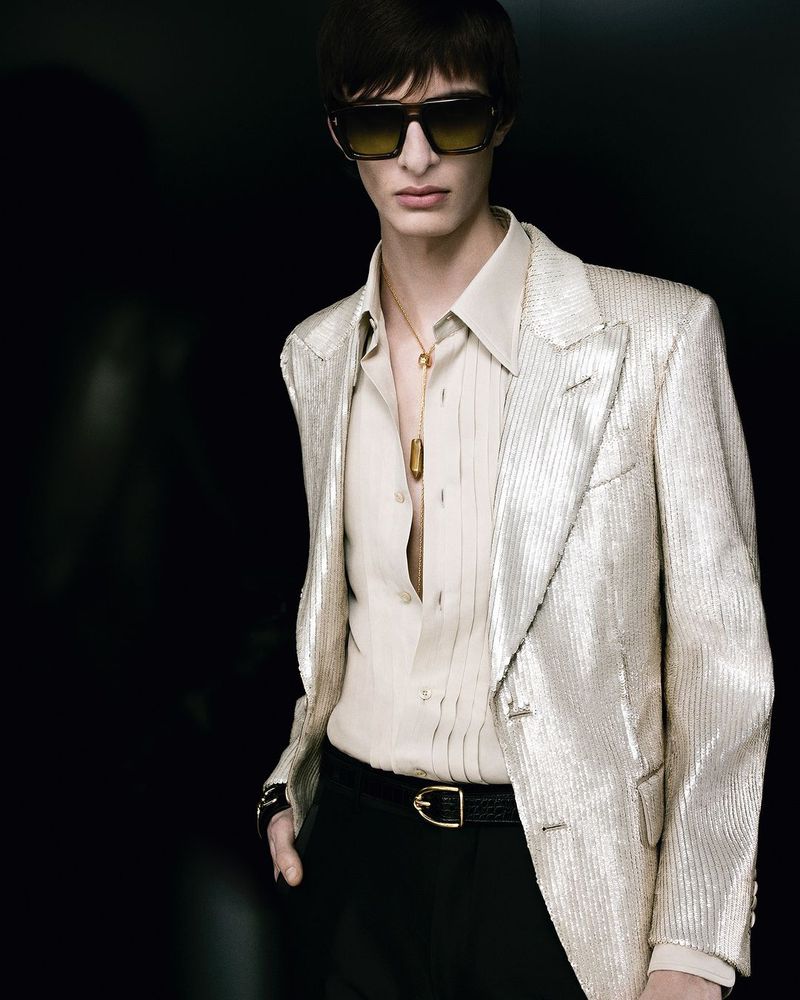 Model wearing a metallic blazer, light shirt, sunglasses, and a pendant necklace against a dark background