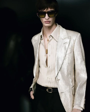 Model wearing a metallic blazer, light shirt, sunglasses, and a pendant necklace against a dark background