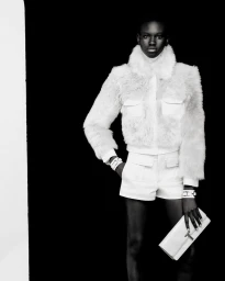 A fashion model posing in a white fur jacket and matching shorts while holding a white clutch against a black background.