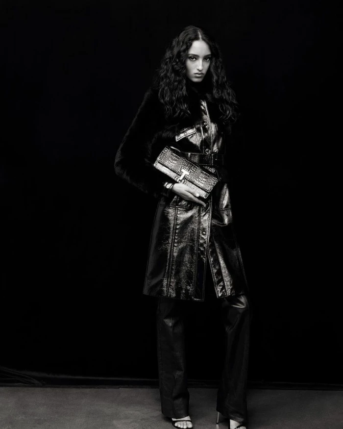 Model Mona Tougaard posing in a belted glossy leather coat, matching trousers, and holding a textured clutch bag against a black backdrop.