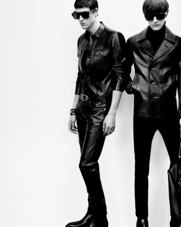 Two male models in black leather jackets, trousers, and dark sunglasses posing against a white seamless background.