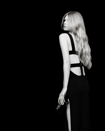 Model looking over her shoulder wearing a black backless cut-out evening gown and holding a clutch against a black background.