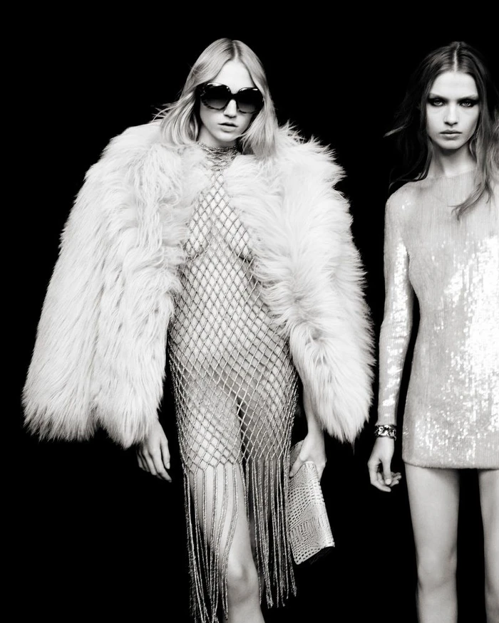 Two models posing against a black background, one wearing a faux fur coat over a metallic mesh dress and sunglasses.