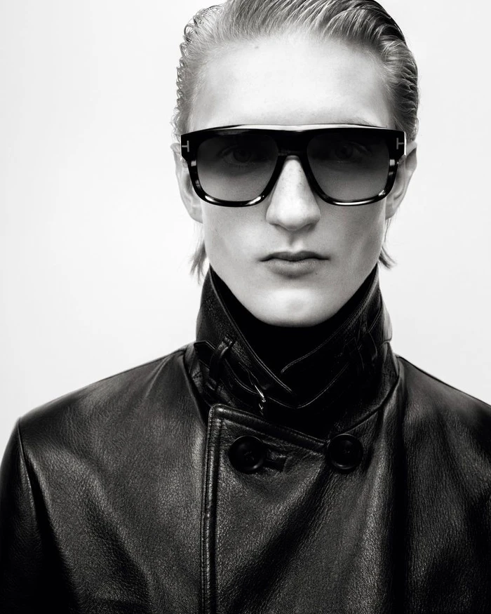Model wearing oversized black sunglasses and a high-collar black leather coat against a white studio background for Tom Ford.