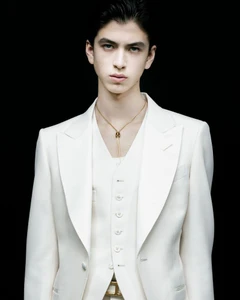 Model wearing an ivory tailored three-piece suit and gold tassel necklace against a solid black background.