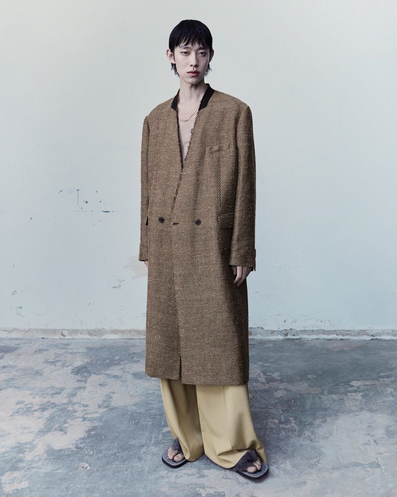 An Asian adult male stands in a photo studio wearing a long brown herringbone jacket, light yellow pants, and sandals.