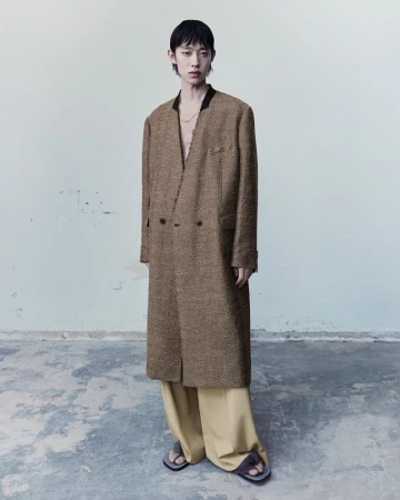 An Asian adult male stands in a photo studio wearing a long brown herringbone jacket, light yellow pants, and sandals.