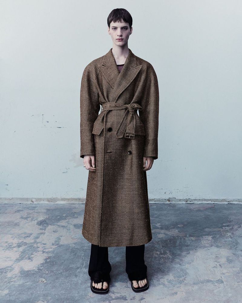 Fashionable adult in a herringbone trench coat with a tie waist standing indoors against a light wall, complemented by dark trousers and sandals.