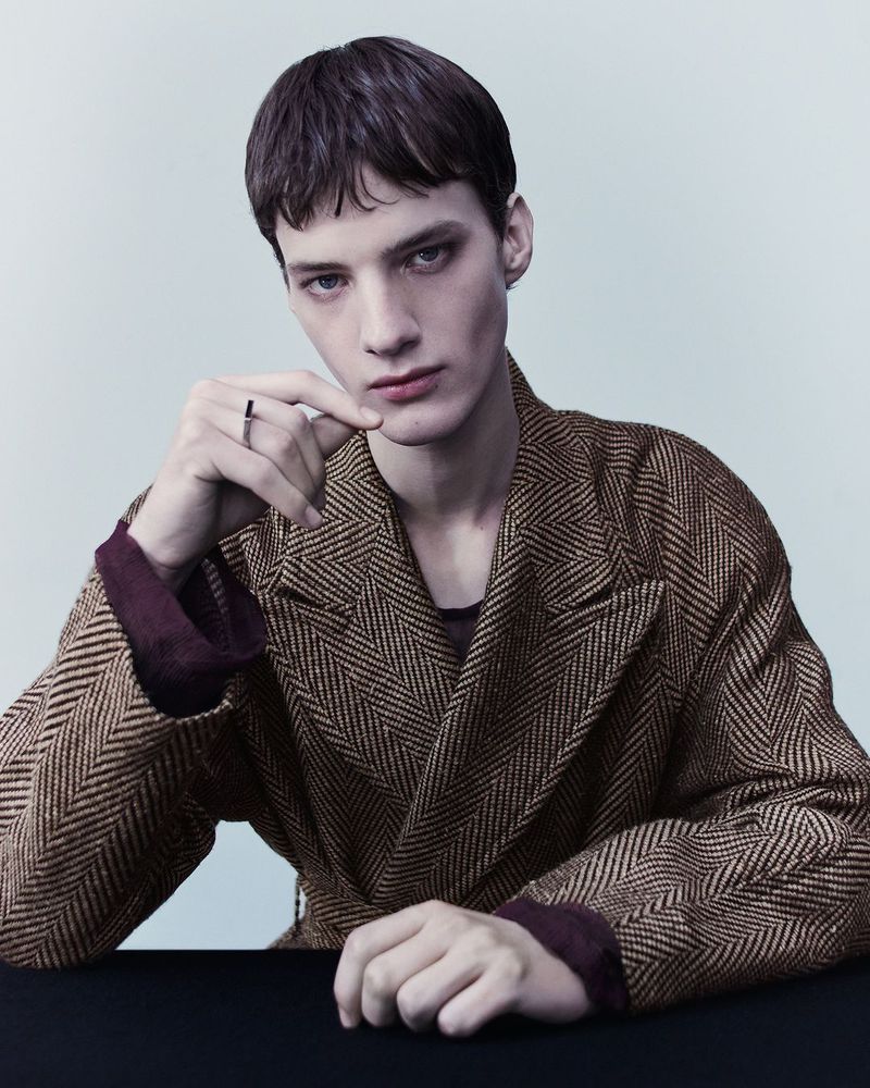 Headshot of a young adult male model wearing a brown herringbone coat and a ring while posing indoors against a light grey background.