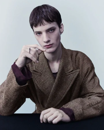 Headshot of a young adult male model wearing a brown herringbone coat and a ring while posing indoors against a light grey background.