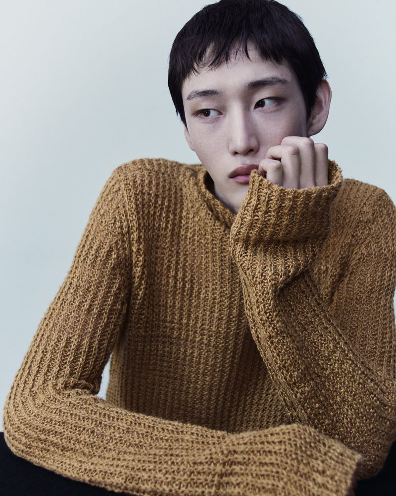 Asian man in a knitted sweater gazes off-camera with a contemplative expression in a studio setting with neutral background lighting.