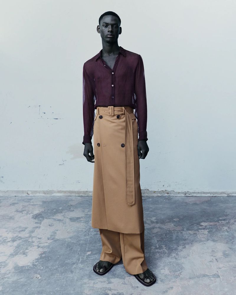 Full-length shot of a Black male model in a semi-sheer shirt and wrap skirt-pants standing against a neutral studio background. 