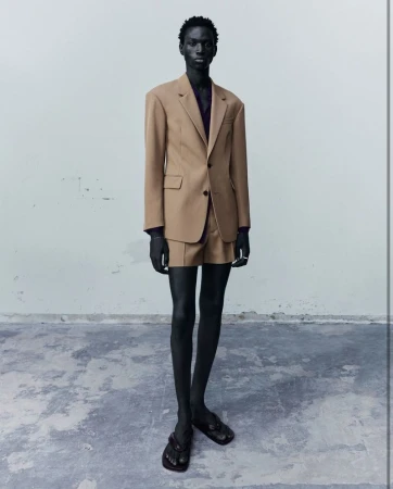 Fashion portrait of a Black male model in a tan suit with shorts, standing in a neutral studio setting. He is wearing sandals.