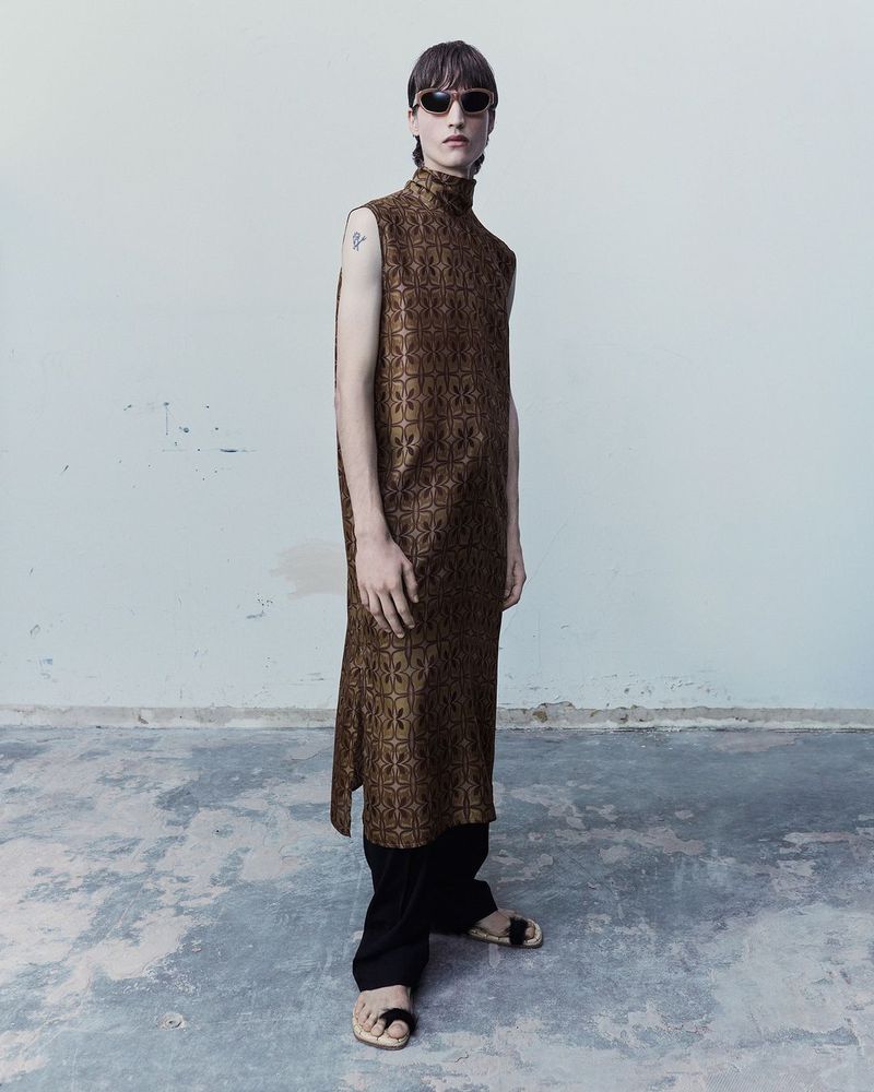 Full shot of a young adult in a sleeveless, patterned tunic, dark pants, and sunglasses, standing in front of a textured white wall.