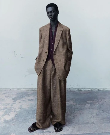 Adult male poses in a herringbone suit in a studio. He wears a dark purple shirt and black sandals.