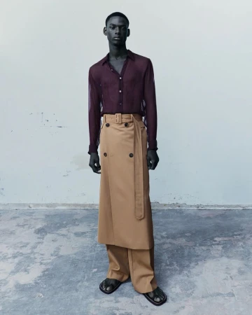 Black male model wearing a sheer burgundy shirt with a camel wrap skirt layered over trousers against a neutral studio backdrop.