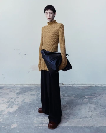 Model wearing a textured mustard yellow knit sweater and wide-leg black trousers holding a large black leather bag.