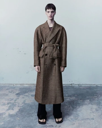 Model wearing an oversized brown textured belted coat and black trousers against a neutral wall for Dries Van Noten S/S 24.