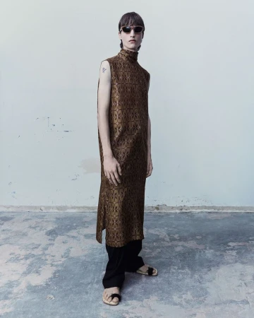 Model in sunglasses wearing a high-neck patterned brown sleeveless tunic over wide black trousers against a neutral wall.