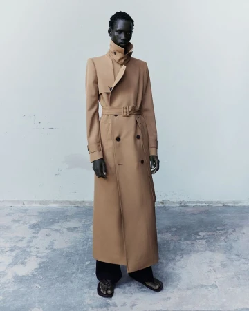 A Black male model stands wearing a full-length belted camel trench coat with a high collar and black trousers in a studio.