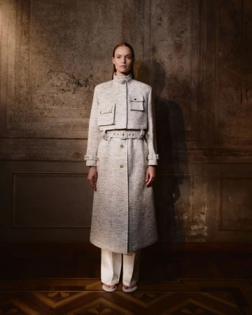Woman in tweed coat with belt and pockets posing against a textured wall with herringbone floor. Full-length fashion portrait.