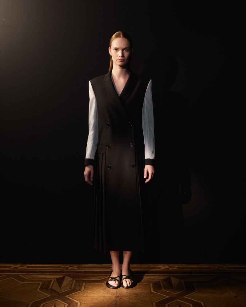 Full-length portrait of a woman in a black jacket dress with white sleeves on a wood floor against a dark background in moody lighting.