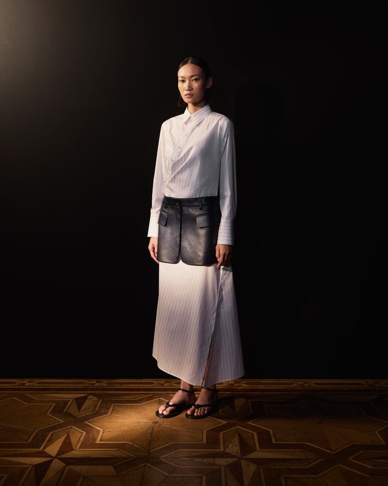 Asian woman in a stylish pinstripe dress with a leather vest stands in a photo studio with warm lighting on a patterned floor.