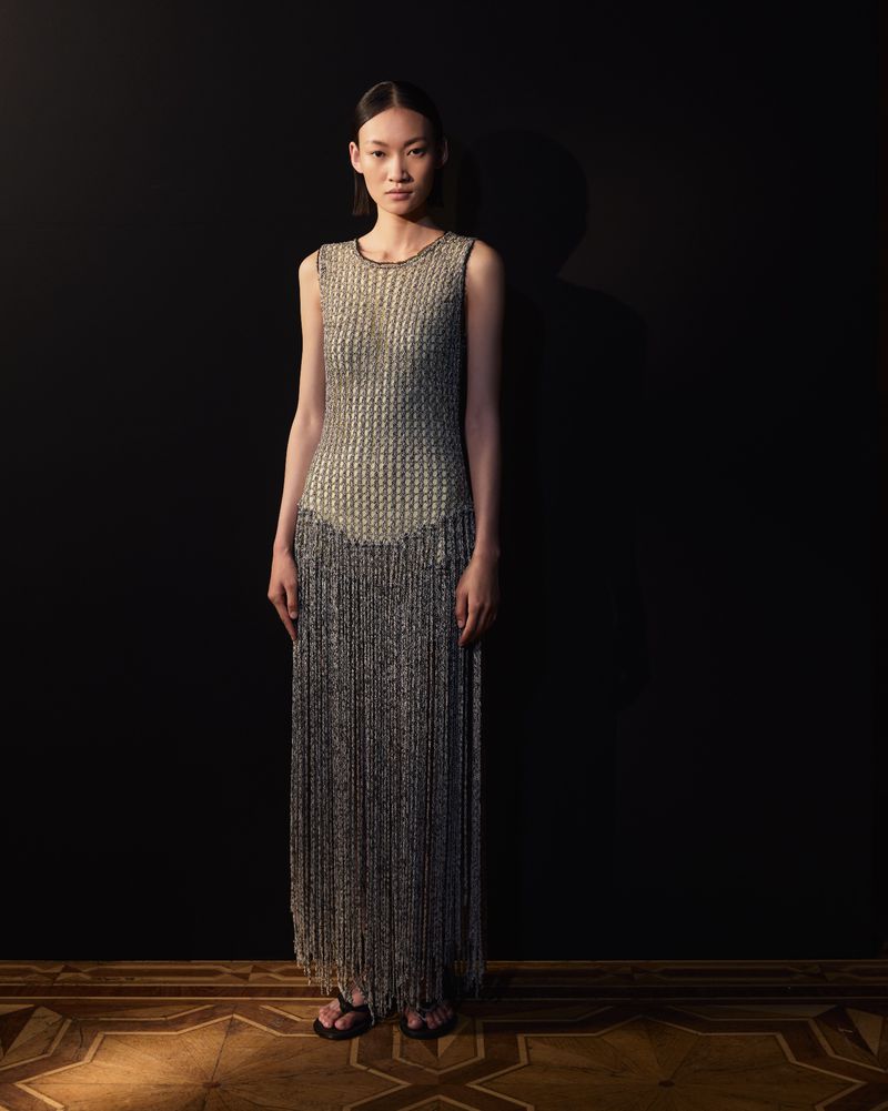 Full-length shot of an adult woman in a sleeveless fringe dress, posing against a dark background on a parquet floor in soft studio lighting.