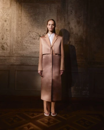 A slender woman poses in a light brown coat and pants, complemented by white sandals, against a textured neutral backdrop.