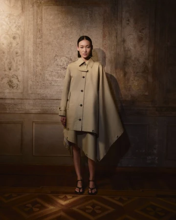 A stylish Asian woman in a beige trench coat poses in front of a textured wall, illuminated by soft, directional lighting and a patterned floor.