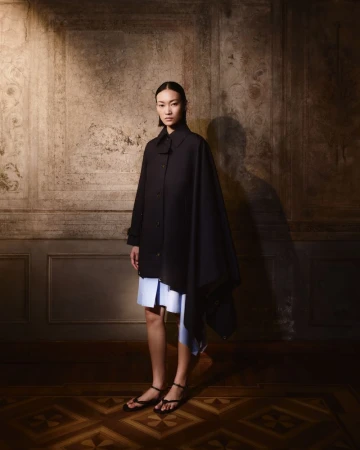 Elegant woman posing in a black coat and light blue dress against a patterned wall and floor for a fashion editorial shoot.