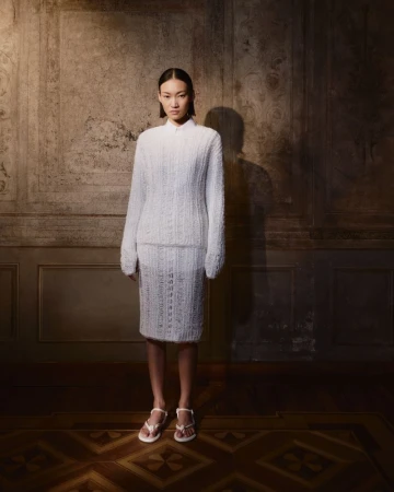 An Asian woman is in a full shot, wearing a knitted skirt and sweater with white sandals, posing in front of a patterned wall.