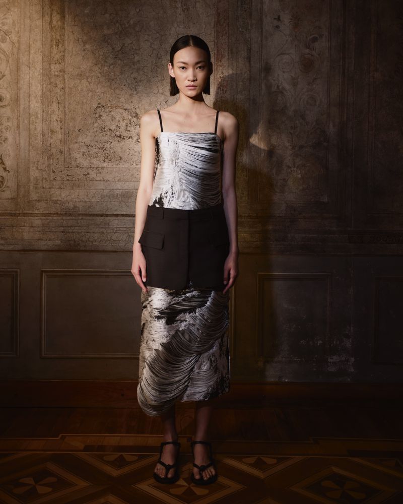 Full shot of a woman in a sleeveless, patterned dress and heels posing in front of an ornate wall in a dimly lit room.
