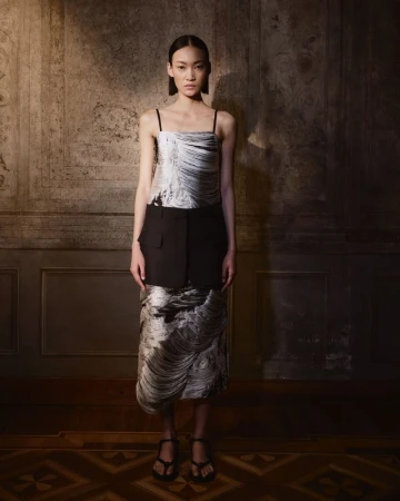 Full shot of a woman in a sleeveless, patterned dress and heels posing in front of an ornate wall in a dimly lit room.